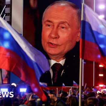 Vladimir Putin on a large screen as a crowd waving Russian flags watch on