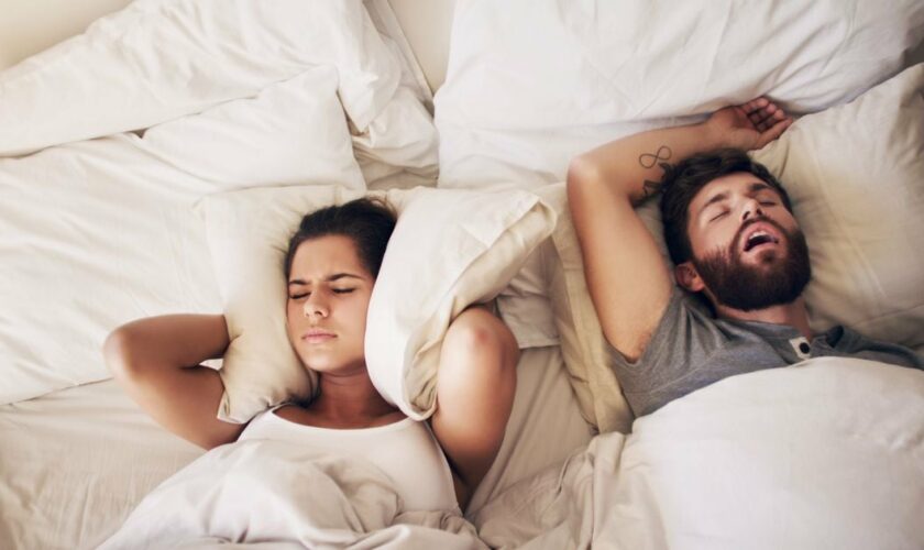 Une jeune femme se bouche les oreilles avec un coussin pendant que son mari ronfle dans le lit.