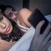 Paar liegt im Bett, sie sieht auf ihr Handy, er blickt misstrauisch hinüber zu ihr
