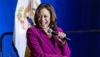 Kamala Harris sitzt auf einer Bühne und lacht, während sie mit links ein Mikro hält